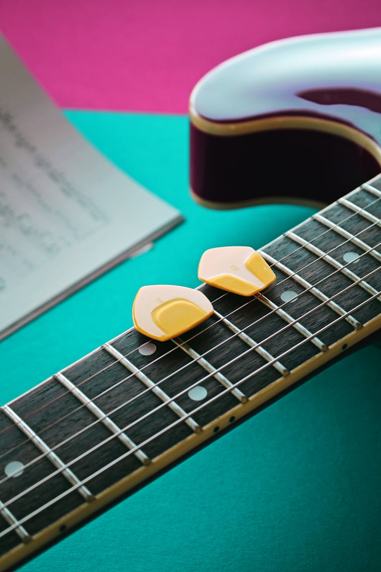 A detailed still life of a guitar fretboard with yellow picks and sheet music in the background.